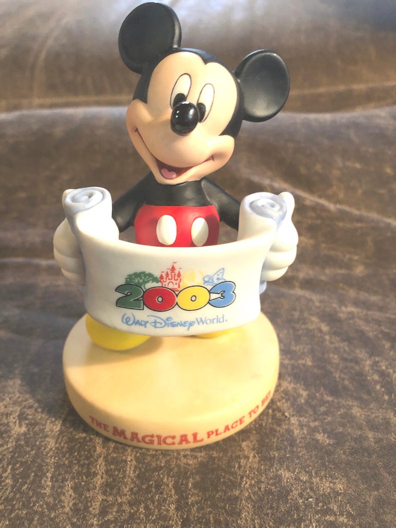 Puede incluir: Una figurita de cer&aacute;mica de Mickey Mouse sosteniendo un rollo con el texto "2003 Walt Disney World. The Magical Place To Be." El rollo es blanco con detalles coloridos y la base es beige.