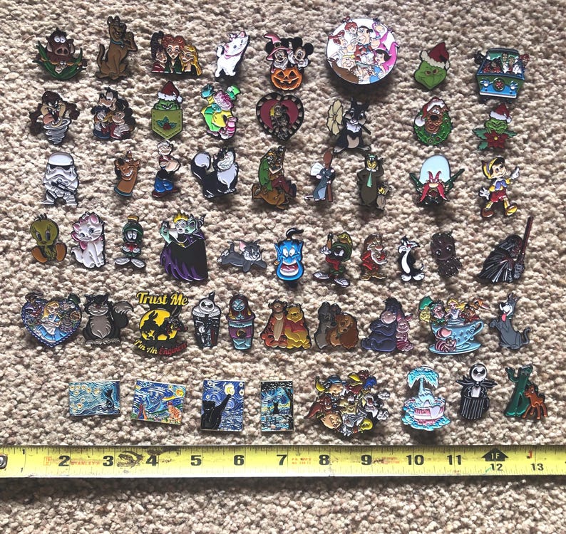 May include: A collection of 50 colorful enamel pins featuring various cartoon characters, including Mickey Mouse, Donald Duck, Goofy, and others. The pins are arranged in rows on a brown carpet-like surface.