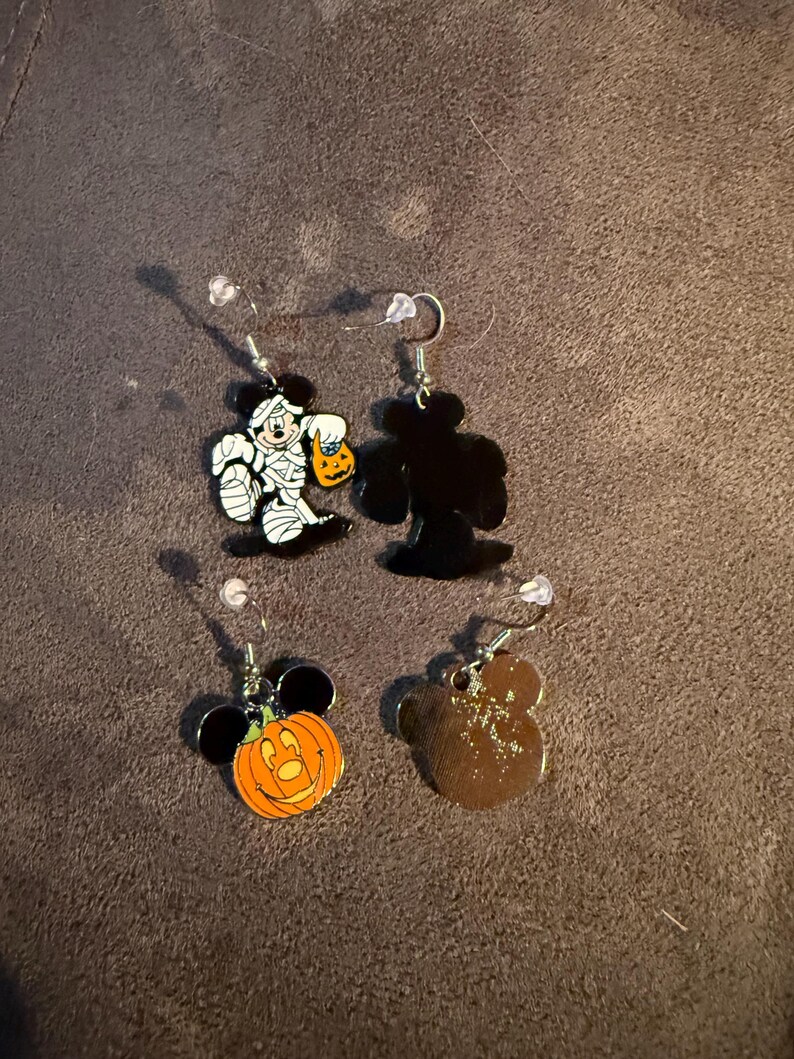 以下が含まれることがあります： ハロウィーンをテーマにした4つのイヤリング。1つのイヤリングは、カボチャのランタンを持っているミイラキャラクターが特徴です。もう1つはキャラクターの黒いシルエットです。3つ目はキャラクターの耳が付いたカボチャです。4つ目は茶色のキャラクターシルエットです。