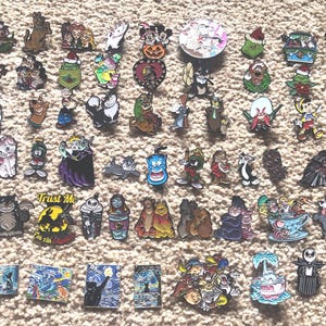 May include: A collection of over 50 colorful enamel pins featuring various cartoon characters, including Mickey Mouse, Bugs Bunny, and Tweety Bird. The pins are arranged in rows on a brown carpet-like surface.