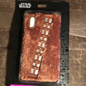 May include: A brown, furry iPhone X/XS phone case resembling Chewbacca from Star Wars. The case features a brown and silver band across the front and is packaged in a black cardboard backing with the Star Wars logo.