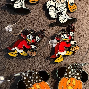 以下が含まれることがあります： ハロウィーンをテーマにしたイヤリング。ミッキーマウスはミイラ、ドナルドダックは悪魔、ミニーマウスはカボチャです。各イヤリングは、銀色のフックが付いたカラフルなエナメルデザインです。