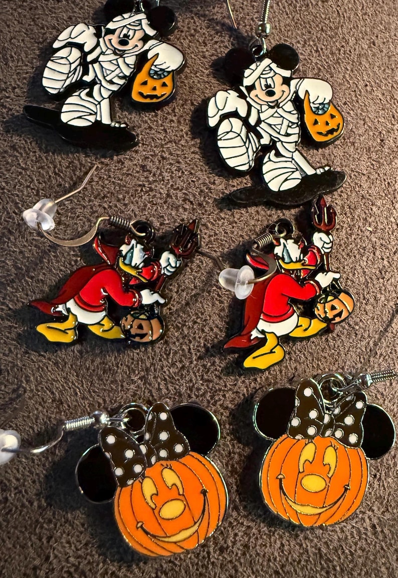 以下が含まれることがあります： ハロウィーンをテーマにしたイヤリング。ミッキーマウスはミイラ、ドナルドダックは悪魔、ミニーマウスはカボチャです。各イヤリングは、銀色のフックが付いたカラフルなエナメルデザインです。