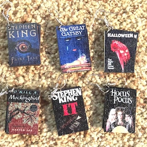 May include: Six book-themed earrings. The earrings feature miniature book covers with titles like "Fairy Tale," "The Great Gatsby," "Halloween," "To Kill a Mockingbird," "IT," and "Hocus Pocus." Each earring has a silver hook.