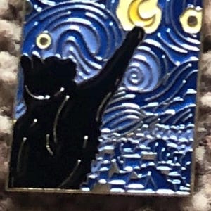May include: A black cat silhouette reaching up towards a yellow crescent moon in a blue and white starry night sky. The image is a pin with a silver border.