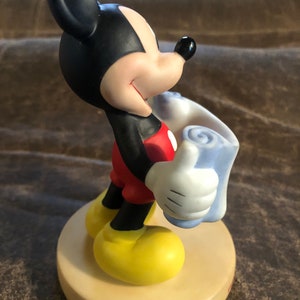 Puede incluir: Una figurita de cer&aacute;mica de Mickey Mouse sosteniendo una manta azul. Mickey lleva pantalones cortos rojos y zapatos amarillos. Est&aacute; sonriendo y mirando hacia la derecha.