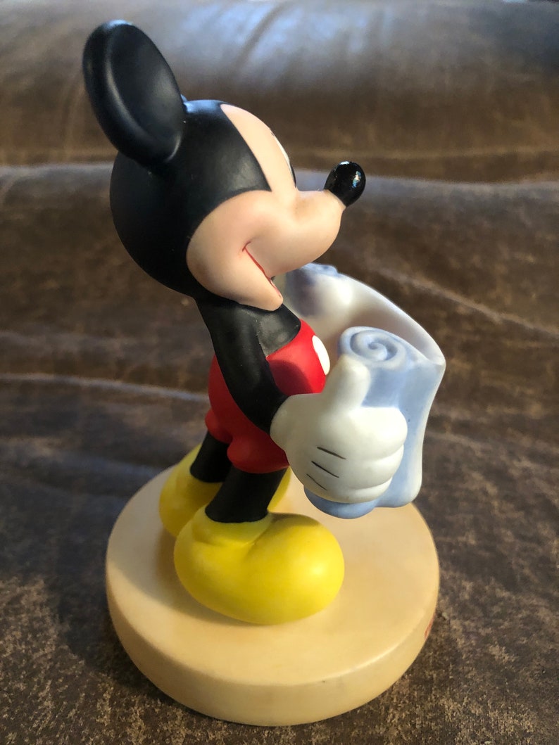 Puede incluir: Una figurita de cer&aacute;mica de Mickey Mouse sosteniendo una manta azul. Mickey lleva pantalones cortos rojos y zapatos amarillos. Est&aacute; sonriendo y mirando hacia la derecha.