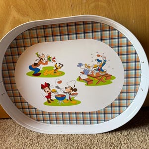 May include: Oval metal serving tray with a white rim and handles. The tray features a plaid pattern and illustrations of Disney characters Goofy, Pluto, Mickey, Minnie, and Donald Duck enjoying a picnic.