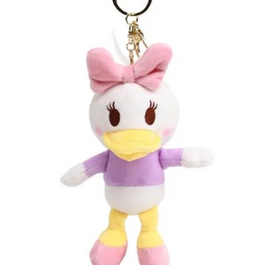 Pode incluir: Um chaveiro de pel&uacute;cia branco em forma de pato de desenho animado, com bico amarelo, camisa roxa e la&ccedil;o rosa.