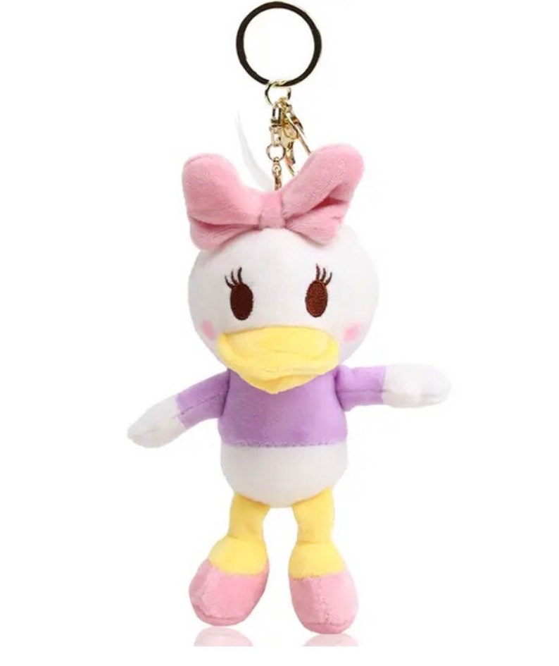 Pode incluir: Um chaveiro de pel&uacute;cia branco em forma de pato de desenho animado, com bico amarelo, camisa roxa e la&ccedil;o rosa.
