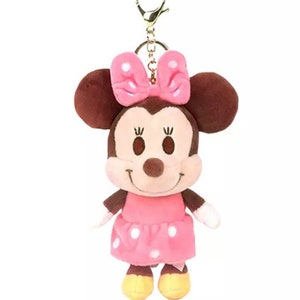 Pode incluir: Um chaveiro de pel&uacute;cia com a Minnie Mouse. Ela est&aacute; usando um vestido rosa com bolinhas brancas e um la&ccedil;o rosa com bolinhas brancas. Ela tem orelhas marrons e um nariz marrom.