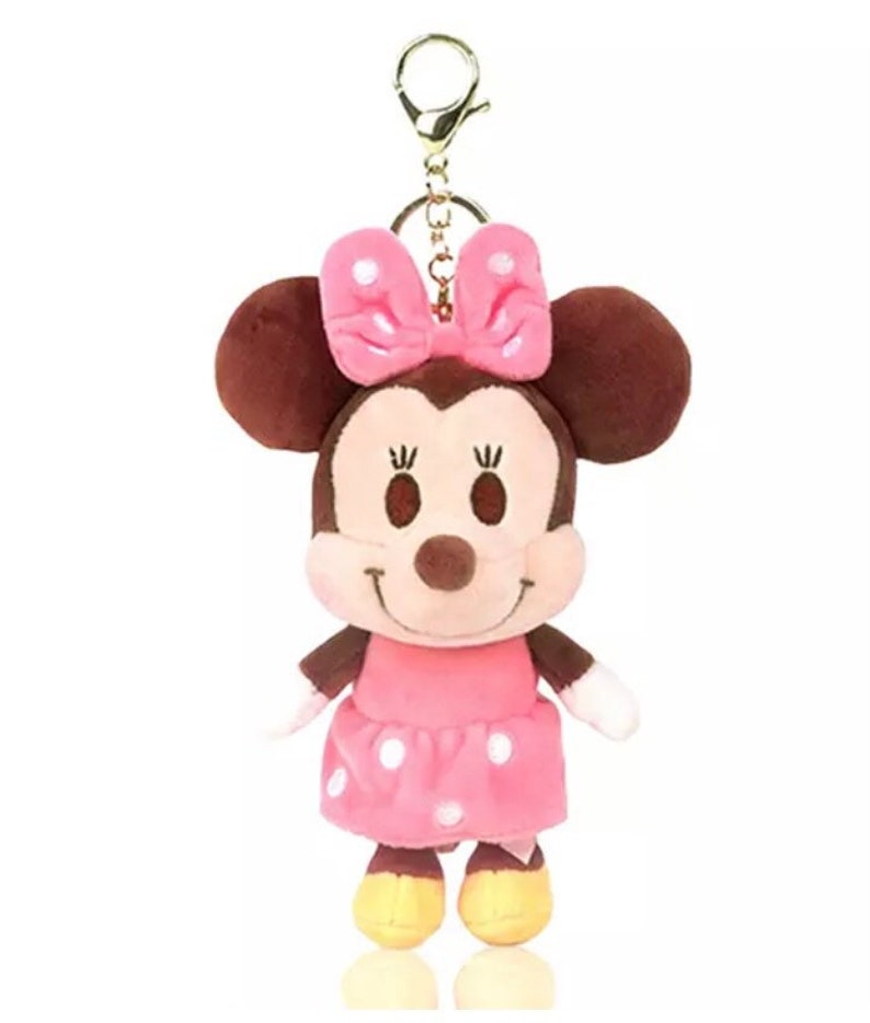 Pode incluir: Um chaveiro de pel&uacute;cia com a Minnie Mouse. Ela est&aacute; usando um vestido rosa com bolinhas brancas e um la&ccedil;o rosa com bolinhas brancas. Ela tem orelhas marrons e um nariz marrom.