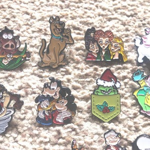 May include: A collection of 11 enamel pins featuring various cartoon characters, including a green Grinch-like figure in a red Santa hat, a brown dog with a blue collar, a white cat with pink ears, and a brown and yellow cartoon character with a red hat.
