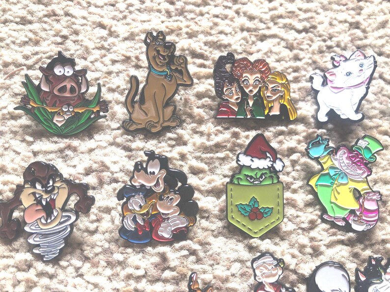 May include: A collection of 11 enamel pins featuring various cartoon characters, including a green Grinch-like figure in a red Santa hat, a brown dog with a blue collar, a white cat with pink ears, and a brown and yellow cartoon character with a red hat.