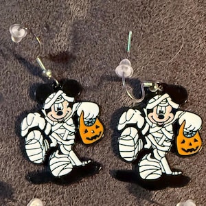 以下が含まれることがあります： ミイラに扮した漫画のキャラクターが特徴のハロウィーンをテーマにしたイヤリングのペア。イヤリングは黒、白、オレンジ色で、ジャック・オー・ランタンのトリートバッグが付いています。イヤリングにはシルバーのフックが付いています。