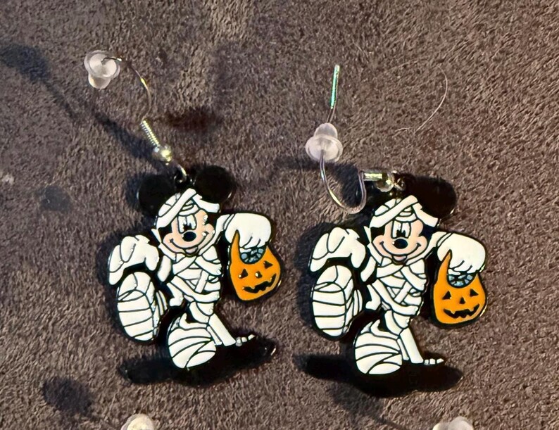 以下が含まれることがあります： ミイラに扮した漫画のキャラクターが特徴のハロウィーンをテーマにしたイヤリングのペア。イヤリングは黒、白、オレンジ色で、ジャック・オー・ランタンのトリートバッグが付いています。イヤリングにはシルバーのフックが付いています。