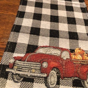 May include: A woven table runner with a black and white checkered pattern. A red vintage truck is embroidered on the runner, its bed filled with pumpkins. The runner is a seasonal autumn decoration.