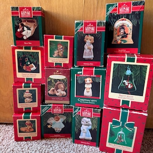 May include: A collection of twelve Hallmark Keepsake Ornament boxes featuring various animal-themed designs. Each box is red and green with a printed image of the specific ornament inside, including cats, bunnies, a lion, a lamb, and a squirrel.