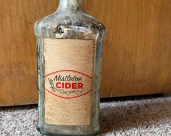 May include: An antique glass bottle with a cork stopper. The bottle has a rectangular label with the words "Mistletoe CIDER" in red and white. The bottle is slightly dirty, suggesting age. The bottle is likely for decorative purposes.