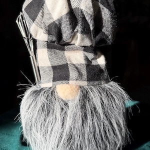 May include: A whimsical gnome figurine wearing a black and white checkered chef's hat and a long, gray beard. A stainless steel whisk is attached to the hat. The gnome has a tan nose and is set against a dark background.