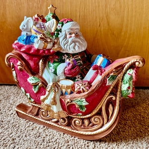 May include: A ceramic Santa Claus figurine in a red sleigh, laden with gifts and a small white rabbit. The sleigh has gold accents and is decorated with holly. A Christmas tree sits atop the gifts.