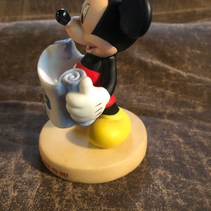 Puede incluir: Una figurita de cer&aacute;mica de Mickey Mouse sosteniendo un rollo. Lleva pantalones cortos rojos y zapatos amarillos. La base de la figurita es beige y tiene las palabras "Back to Be" escritas en ella.