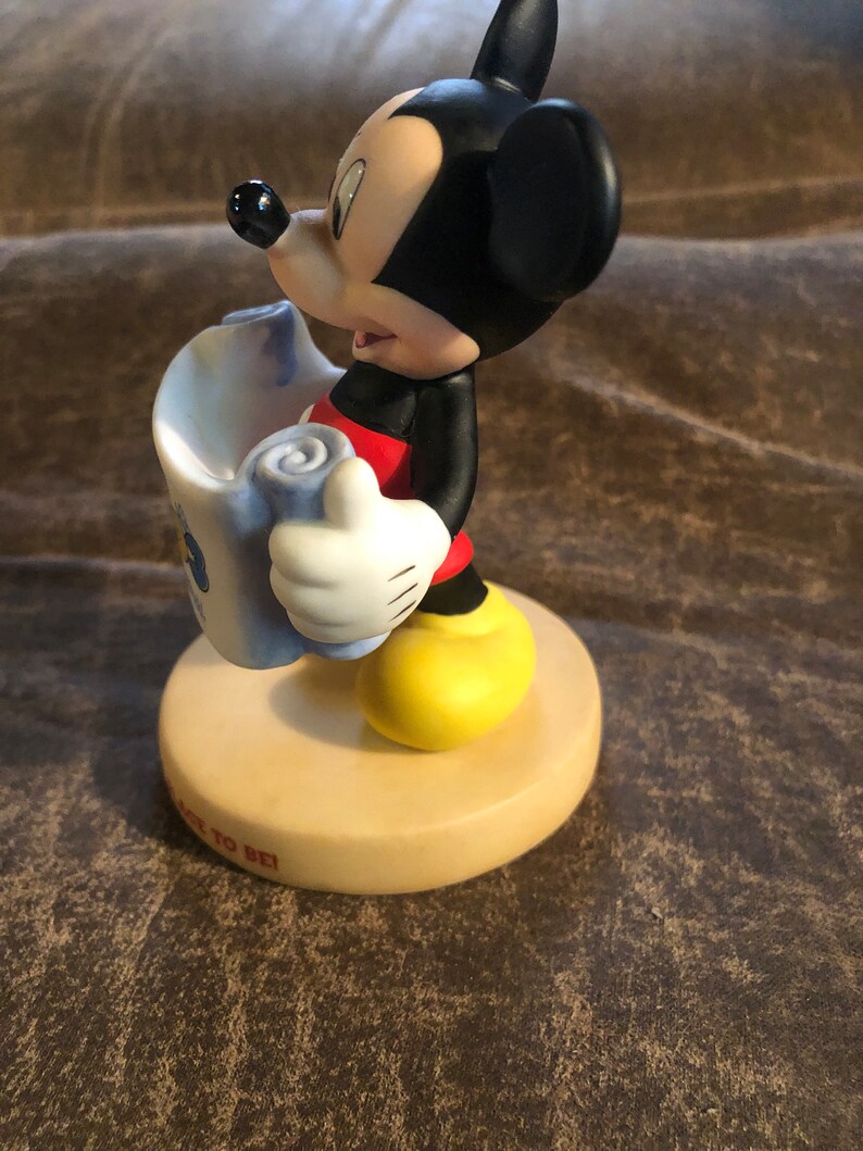 Puede incluir: Una figurita de cer&aacute;mica de Mickey Mouse sosteniendo un rollo. Lleva pantalones cortos rojos y zapatos amarillos. La base de la figurita es beige y tiene las palabras "Back to Be" escritas en ella.