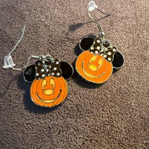 以下が含まれることがあります： ハロウィーンをテーマにしたイヤリングのペア。各イヤリングは、黒と白のドット柄のリボンと黒いミッキーマウスの耳が付いた、笑顔のオレンジ色のカボチャが特徴です。イヤリングはシルバーカラーのフィッシュフックのイヤリングワイヤーが付いています。