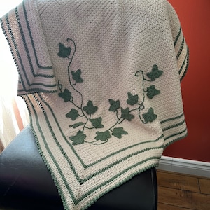May include: A crocheted throw blanket with a green ivy design. The blanket is white with a green border and is folded on a black chair.