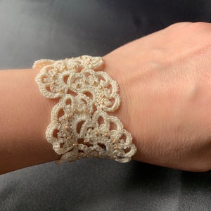 Può includere: Un braccialetto bianco all'uncinetto con perline dorate. Il braccialetto è realizzato con punti delicati e intricati e presenta un design floreale.