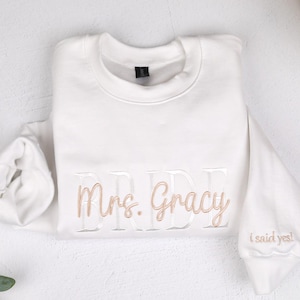 May include: A white sweatshirt with the word "BRIDE" and "Mrs. Gracie" embroidered in a light brown color. The sleeve has the phrase "I said yes!" embroidered in the same color. The sweatshirt is laid flat on a white surface.