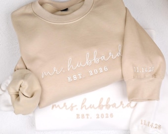 May include: A beige sweatshirt with white embroidery reading mr. hubbard EST. 2026 and 11.14.26 on the sleeve, layered over a white sweatshirt with beige embroidery reading mrs. hubbard EST. 2026 and 11.14.26 on the sleeve, both with crew necks.
