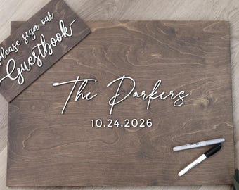 May include: A rectangular dark brown wooden guestbook sign with white 3D lettering that reads The Parkers and the date 10.24.2026. A smaller matching sign reads please sign our Guestbook. Two markers are placed on the main wooden board surface.