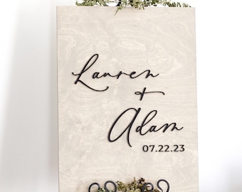 May include: A rectangular sign with a light beige marbled pattern featuring the black script text Lauren + Adam and the date 07.22.23, displayed on a black metal easel decorated with small green sprigs at the top and bottom edges.