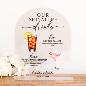 May include: A wedding drink sign with the words "Our Signature Drinks" in elegant script. The sign features illustrations of two cocktails: an Arnold Palmer and a Raspberry Lemon Drop. The names of the couple and the wedding date are also displayed.