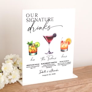 May include: A white acrylic sign with the words "OUR SIGNATURE drinks" in elegant script. Below are watercolor illustrations of three cocktails: Kentucky Mule, Blackberry Martini, and Rum Punch. The sign includes the names of the drinks and ingredients. The names "Isabelle & Alexander" and the date "August 22, 2026" are also printed.