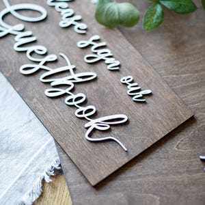 a wooden sign with a cursive message that reads "Please sign our guestbook" placed on a wooden surface.
