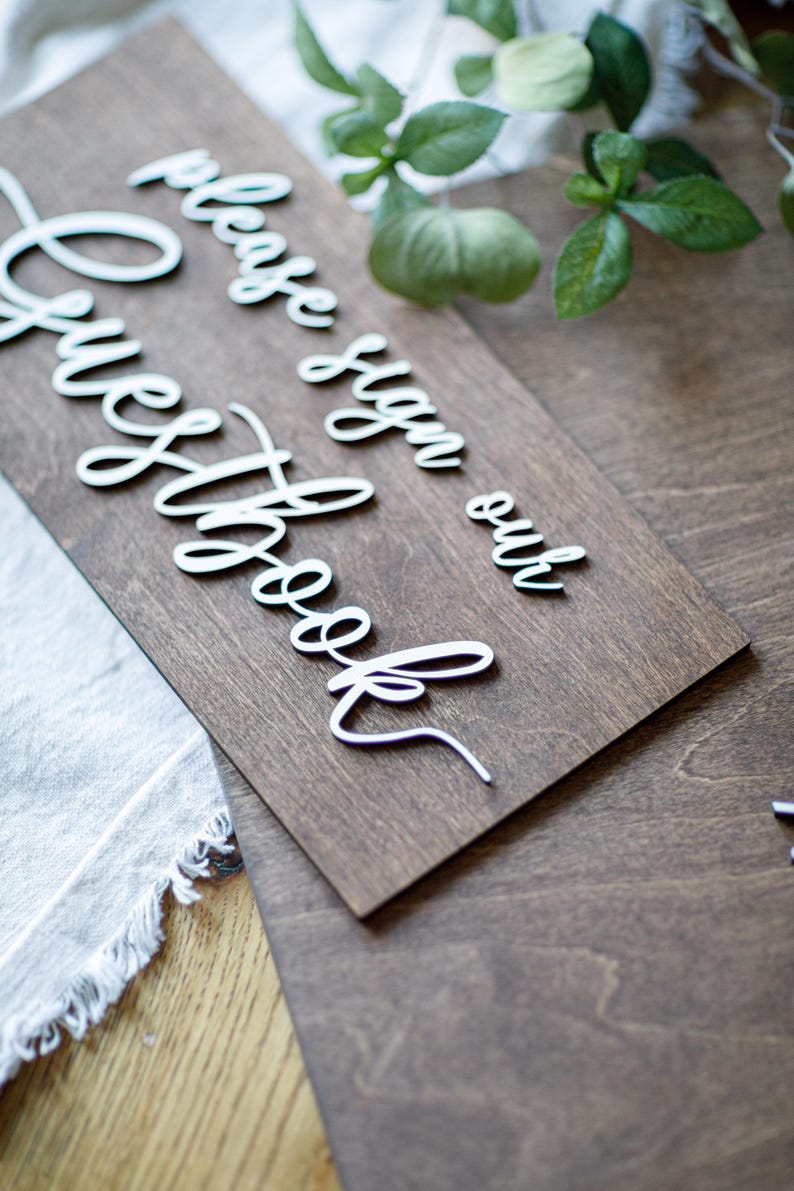 a wooden sign with a cursive message that reads "Please sign our guestbook" placed on a wooden surface.