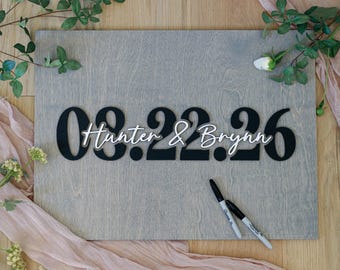 May include: A rectangular gray wooden board featuring large black cutout numbers displaying the date 08.22.26. Overlaid in the center is white cursive text reading Hunter & Brynn. Two black permanent markers are placed on the board surface.