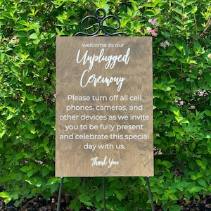 May include: A wooden sign with the text "Welcome to our Unplugged Ceremony" and a message asking guests to turn off their phones and cameras to be fully present for the ceremony. The sign is on a black easel.