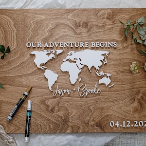 May include: A wooden world map with the text "OUR ADVENTURE BEGINS" and the names "Jason + Brooke" below the map. The date "04.12.2025" is displayed at the bottom right. Two pens are placed on the wooden surface.