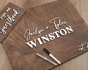 May include: A dark brown wooden guestbook sign with white 3D lettering reading "please sign our Guestbook" and a larger board featuring "Jaclyn + Tyler WINSTON" and "est. 2026" in white, accompanied by one black and one silver permanent marker.