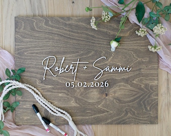May include: A rectangular dark brown wooden guest book sign featuring the white script text Robert + Sammi and the date 05.02.2026 in a serif font. Two black markers are placed on the board, which is decorated with white flowers and pink fabric.