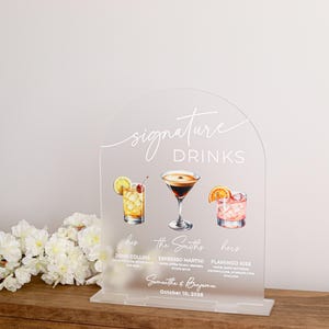 May include: An arched, frosted acrylic sign featuring the text "signature DRINKS" in white. It displays illustrations of three cocktails labeled "John Collins," "Espresso Martini," and "Flamingo Kiss," with the names "Samantha & Benjamin" and "October 10, 2025."