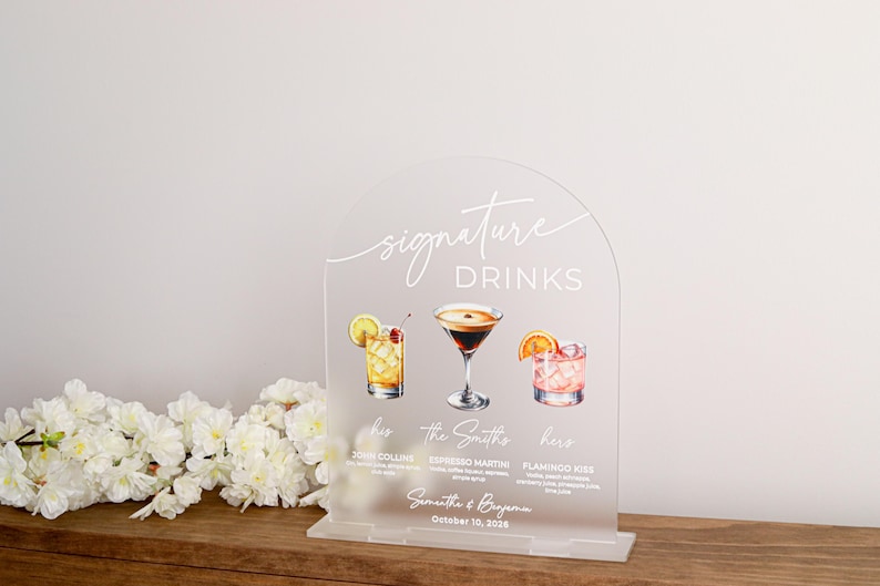 May include: An arched, frosted acrylic sign featuring the text "signature DRINKS" in white. It displays illustrations of three cocktails labeled "John Collins," "Espresso Martini," and "Flamingo Kiss," with the names "Samantha & Benjamin" and "October 10, 2025."