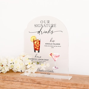 May include: An arched clear acrylic sign featuring the text "OUR SIGNATURE drinks" with illustrations of an Arnold Palmer and a Raspberry Lemon Drop cocktail. The sign lists ingredients for each drink, the names "Madeline & Nicholas", and "June 27, 2026".