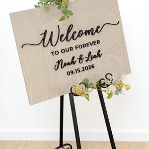 May include: A rectangular light wood sign with black raised lettering that reads Welcome TO OUR FOREVER Noah & Leah 09.15.2026. The sign is displayed on a black metal easel and is decorated with small green leaves and white flowers.