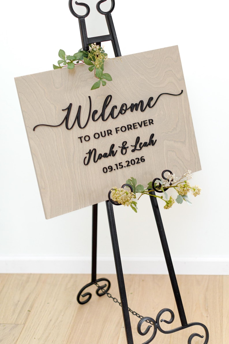 May include: A rectangular light wood sign with black raised lettering that reads Welcome TO OUR FOREVER Noah & Leah 09.15.2026. The sign is displayed on a black metal easel and is decorated with small green leaves and white flowers.