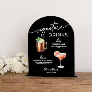 May include: An arched black acrylic sign featuring the text Signature Drinks, his Moscow Mule, and hers Grapefruit Margarita with illustrations of each cocktail. The sign includes the names Olivia & Gabriel and the date May 16, 2026.