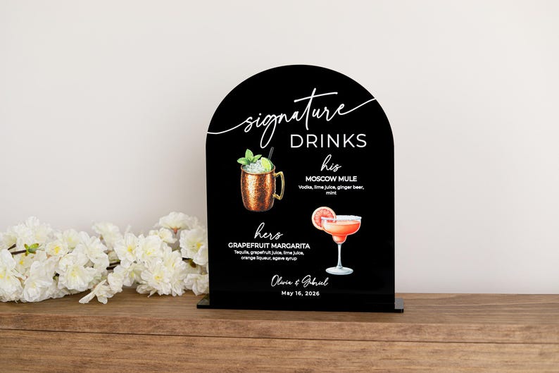 May include: An arched black acrylic sign featuring the text Signature Drinks, his Moscow Mule, and hers Grapefruit Margarita with illustrations of each cocktail. The sign includes the names Olivia & Gabriel and the date May 16, 2026.